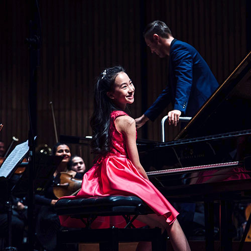 Harmony Zhu seated at a grand piano, orchestra in the background.