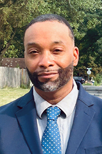 Headshot of Damonni Farley, standing outside, wearing a blue suit, light shirt, blue tie with white dots.
