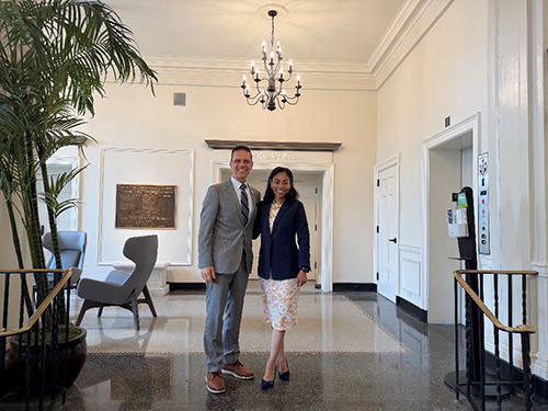 Assemblyman Angelo Santabarbara in a light gray suit and President Dr. Nicole Reaves in a navy jacket over a white floral dress, stand together in the Washington Street Lobby of Eslton Hall.