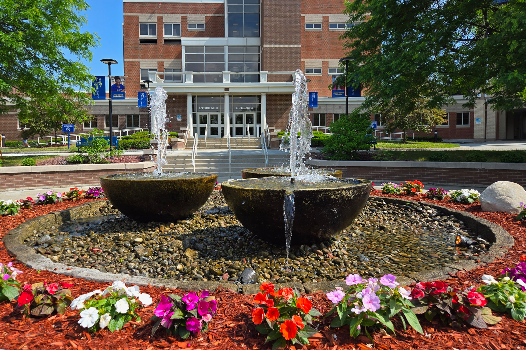 The fountain in the Quad at SUNY Schenectady, surrounded by flowers. Stockade Building in the background.
