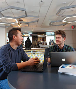 Two students in the modern Begley Learning Commons, talking together at a table with a laptop. 