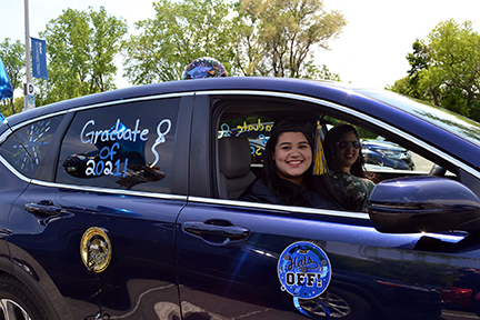 Graduate in cap and gown sitting in a car that is decorated with signs and balloons.