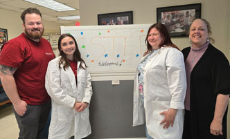 Three CSTEP students and the CSTEP director stand by a "welcome" whiteboard at Albany Med. 