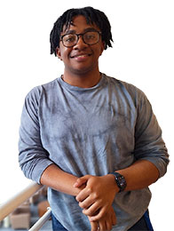 Black male student, wearing glasses and a bluish gray long sleeved shirt, slightly leaning on a banister.