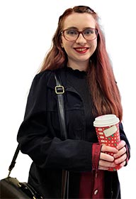 Student with long hair, wearing glasses, holding a coffee cup, standing outside a classroom.
