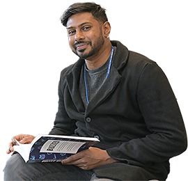 Male student wearing a dark sweater and jeans, seated, reading a book.