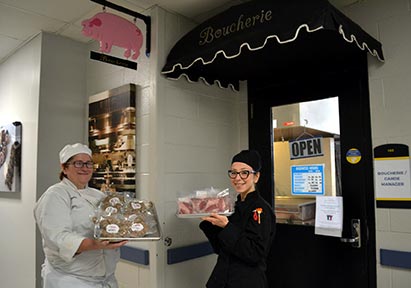 A faculty member and a student holding trays of meat for sale in front of the Boucherie entrance.