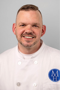 Headshot of Chef Shawn Smith, wearing a white chef coat with the Mazzone Hospitality logo on the left chest.