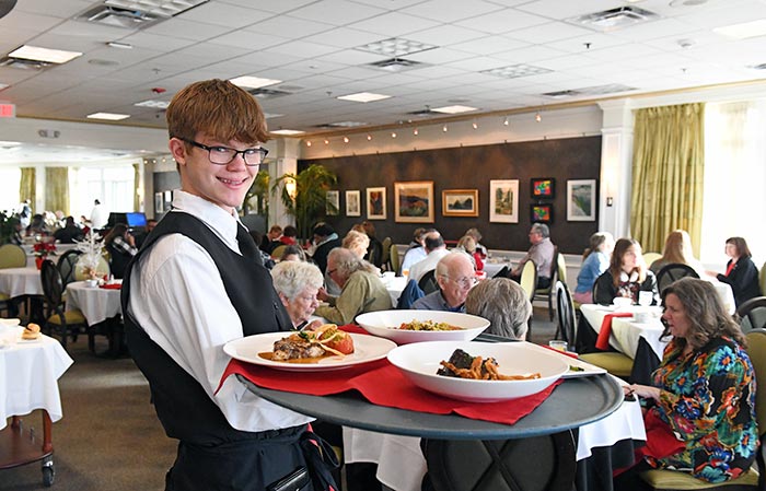 Student server holding a tray of entrees with the Casola Dining Room, full of patrons, behind him.