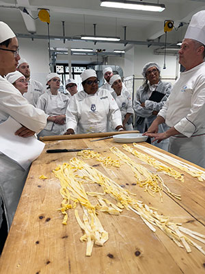 SUNY Schenectady culinary students in their chef whites, stand with an instructor over a wooden table covered with homemade pasta.