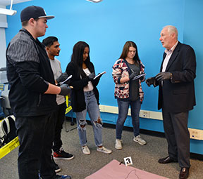 Professor and four students holding clipboards standing over a mock crime scene.