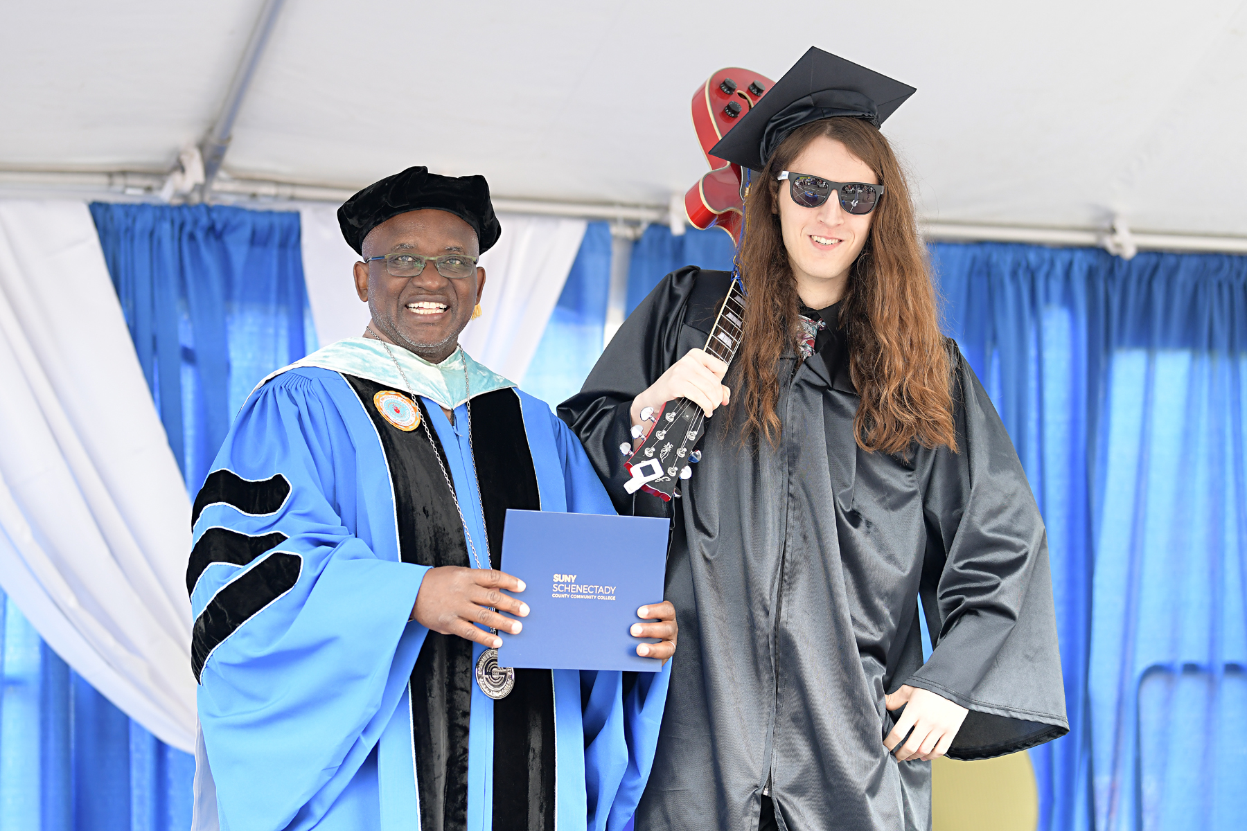 Graduate on stage holding guitar, standing with Dr. Moono