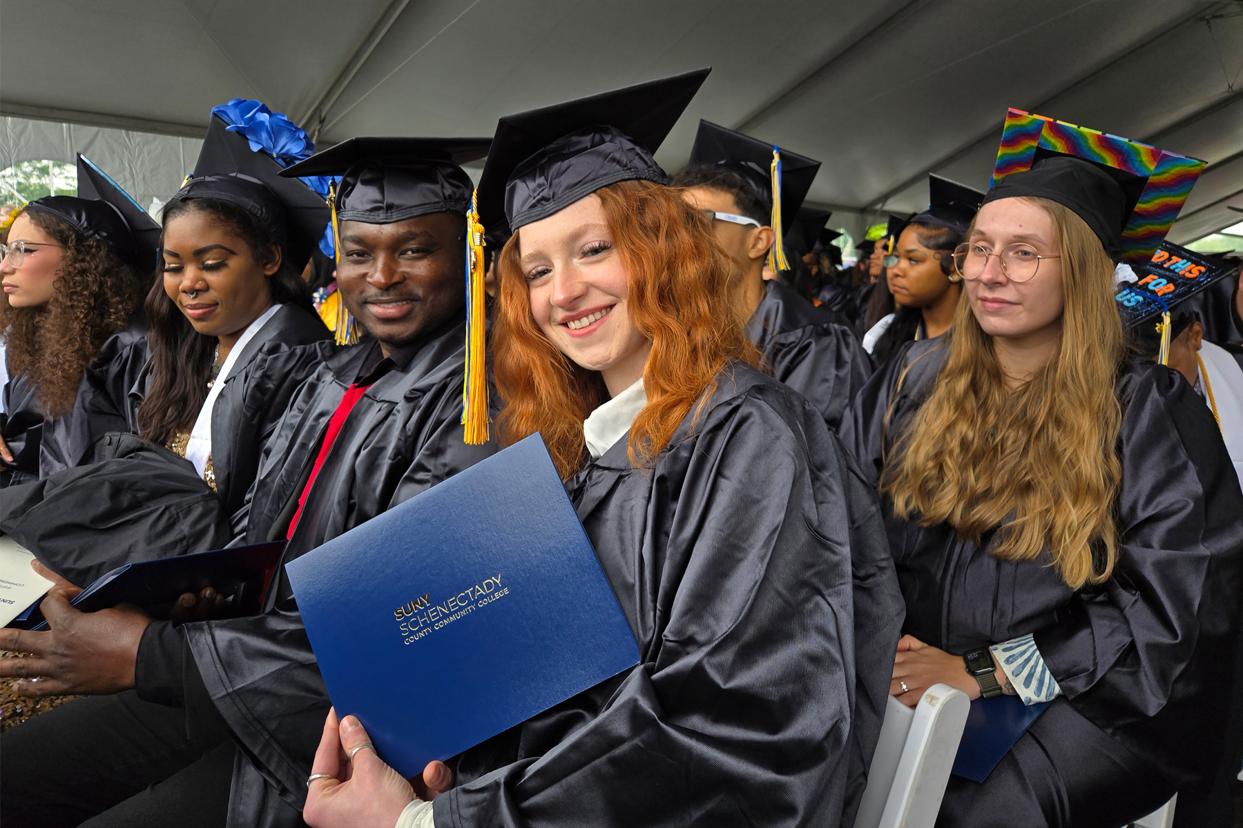Graduates seated, holding degree covers, smiling