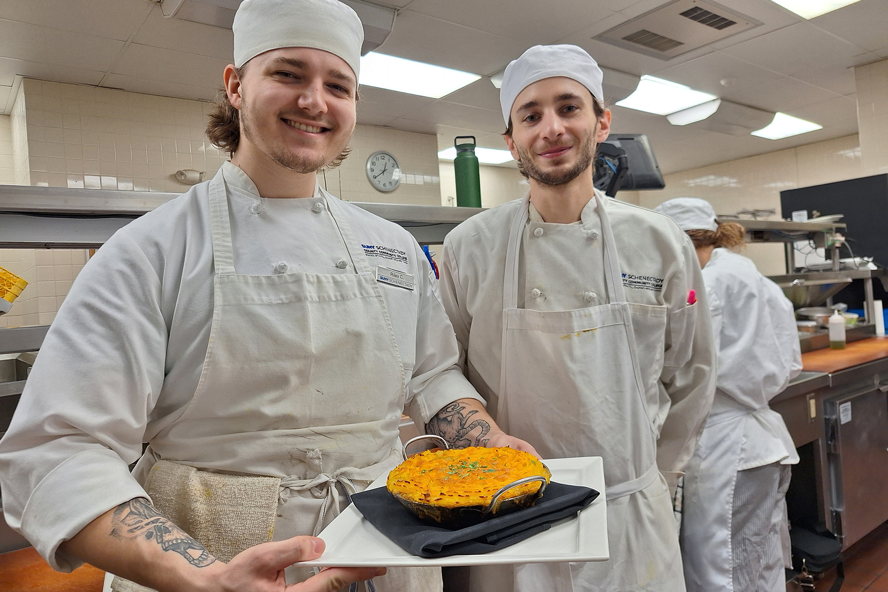 Two students wearing uniforms, standing inside Culinary Lab showing plate of shepard's pie