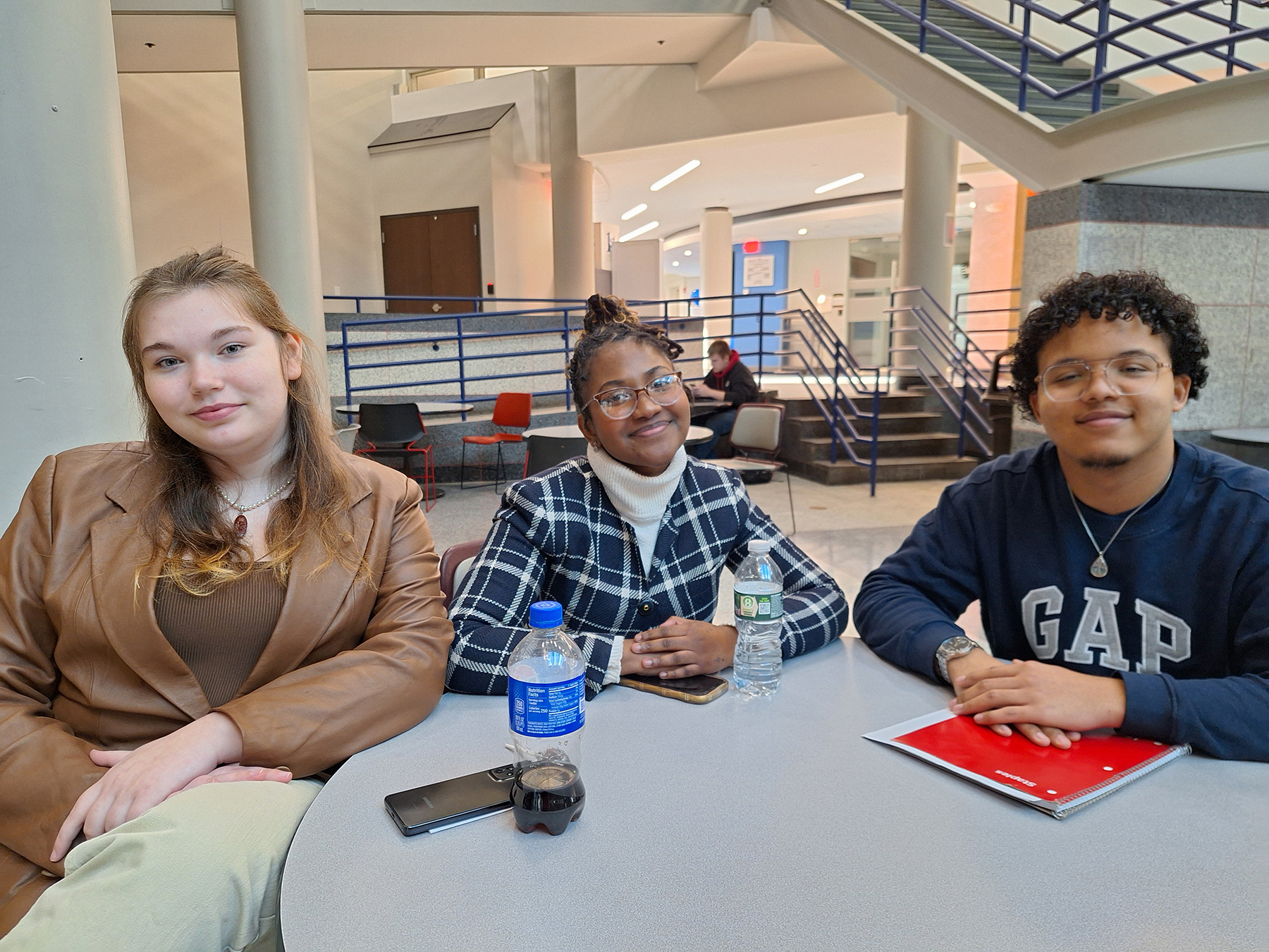 Three students seated at table in cafeteria, smiling