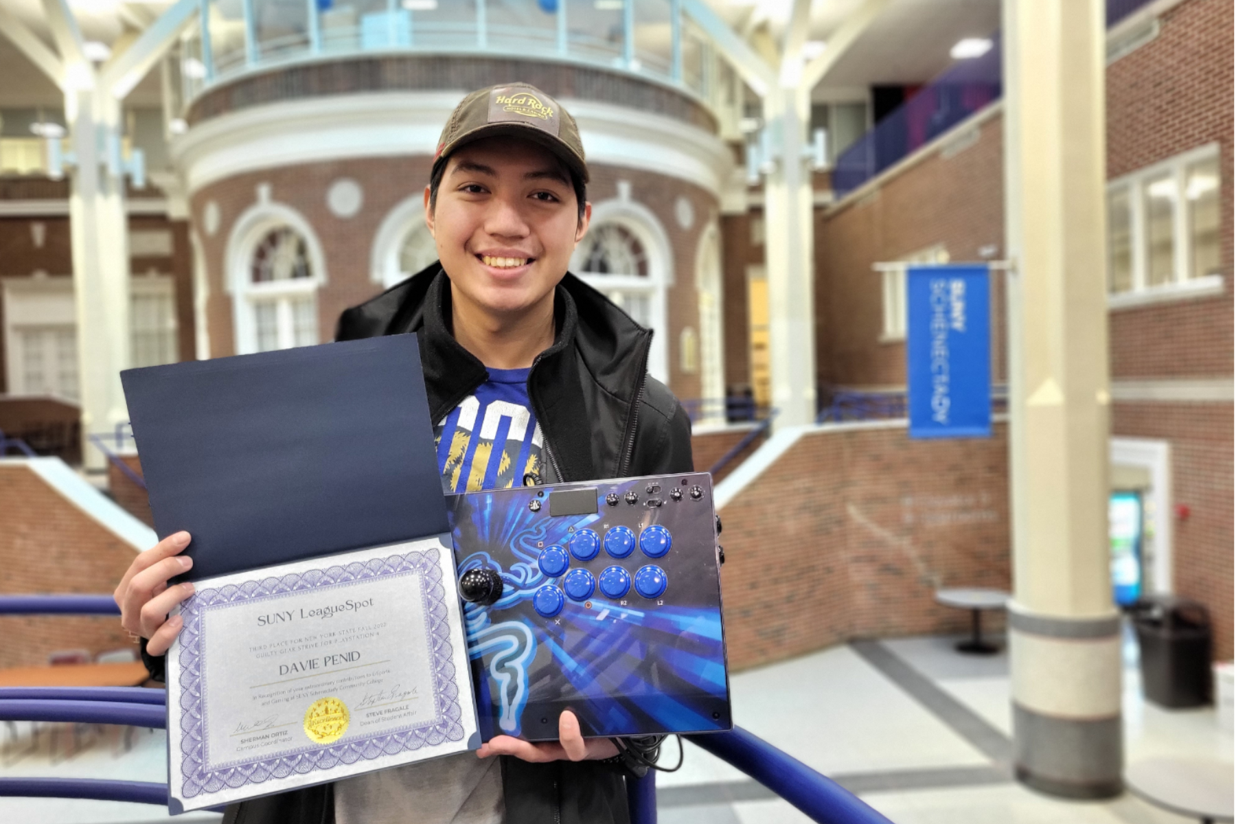 Davie Penid holding e-gamin competition certificate and gaming console, standing on stairs overlooking cafeteria