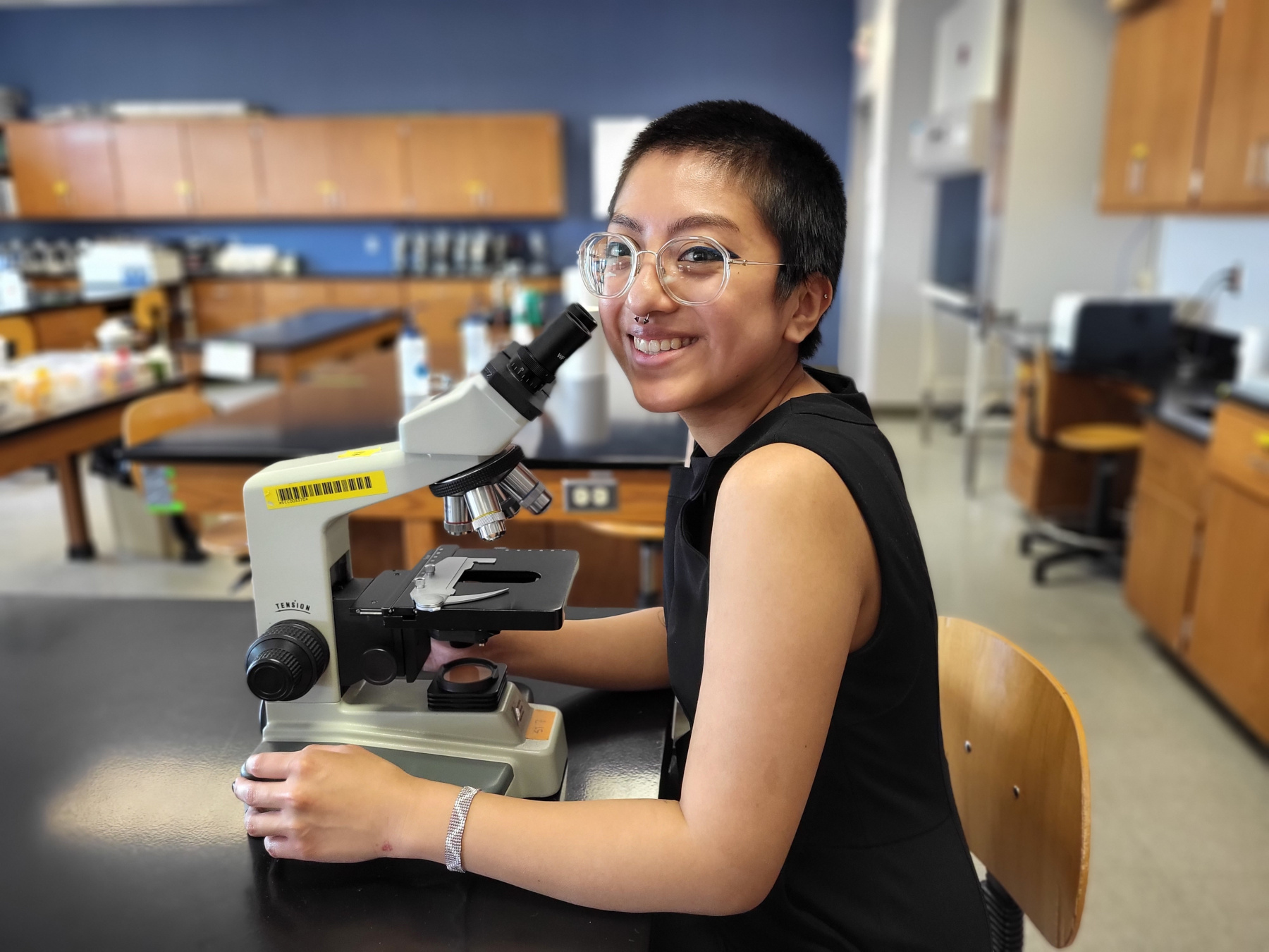 Jennifer Diaz, sitting at microscope in Biology Lab, smiling