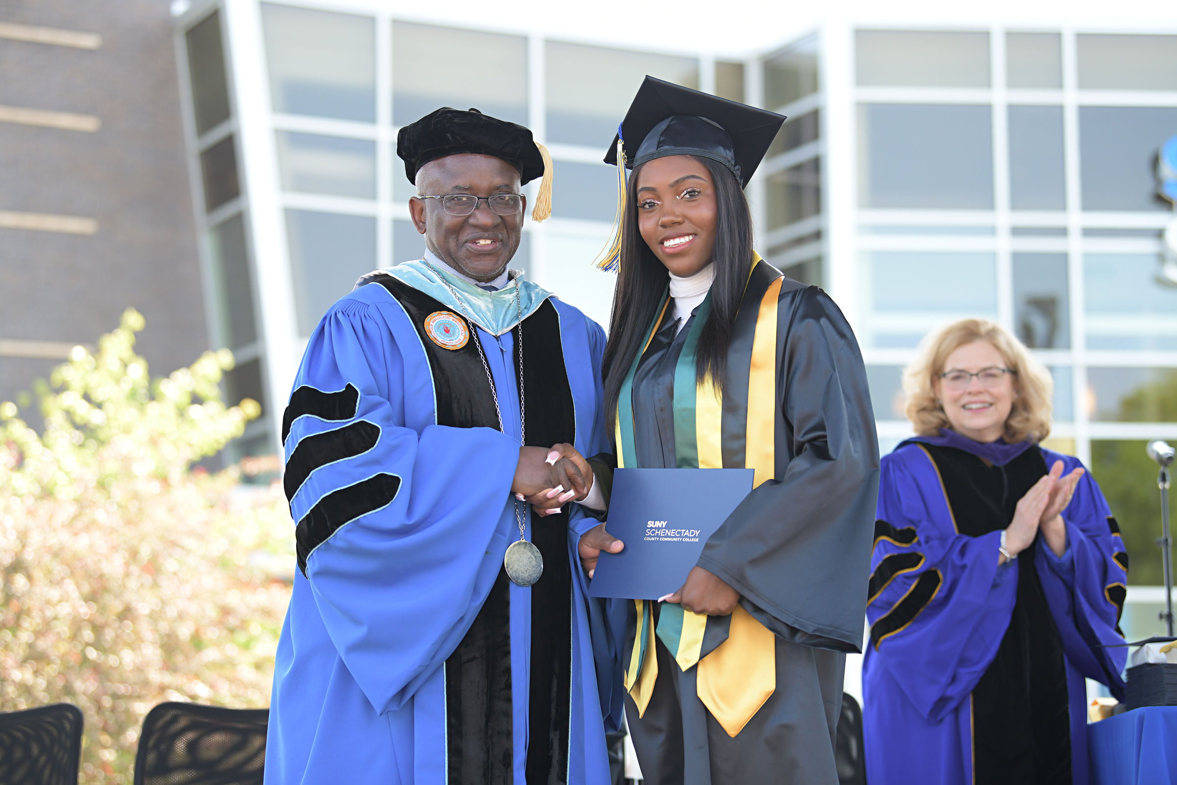 Justina Campbell in cap and gown shaking hands with College President Dr. Moono on stage outside during Commencement