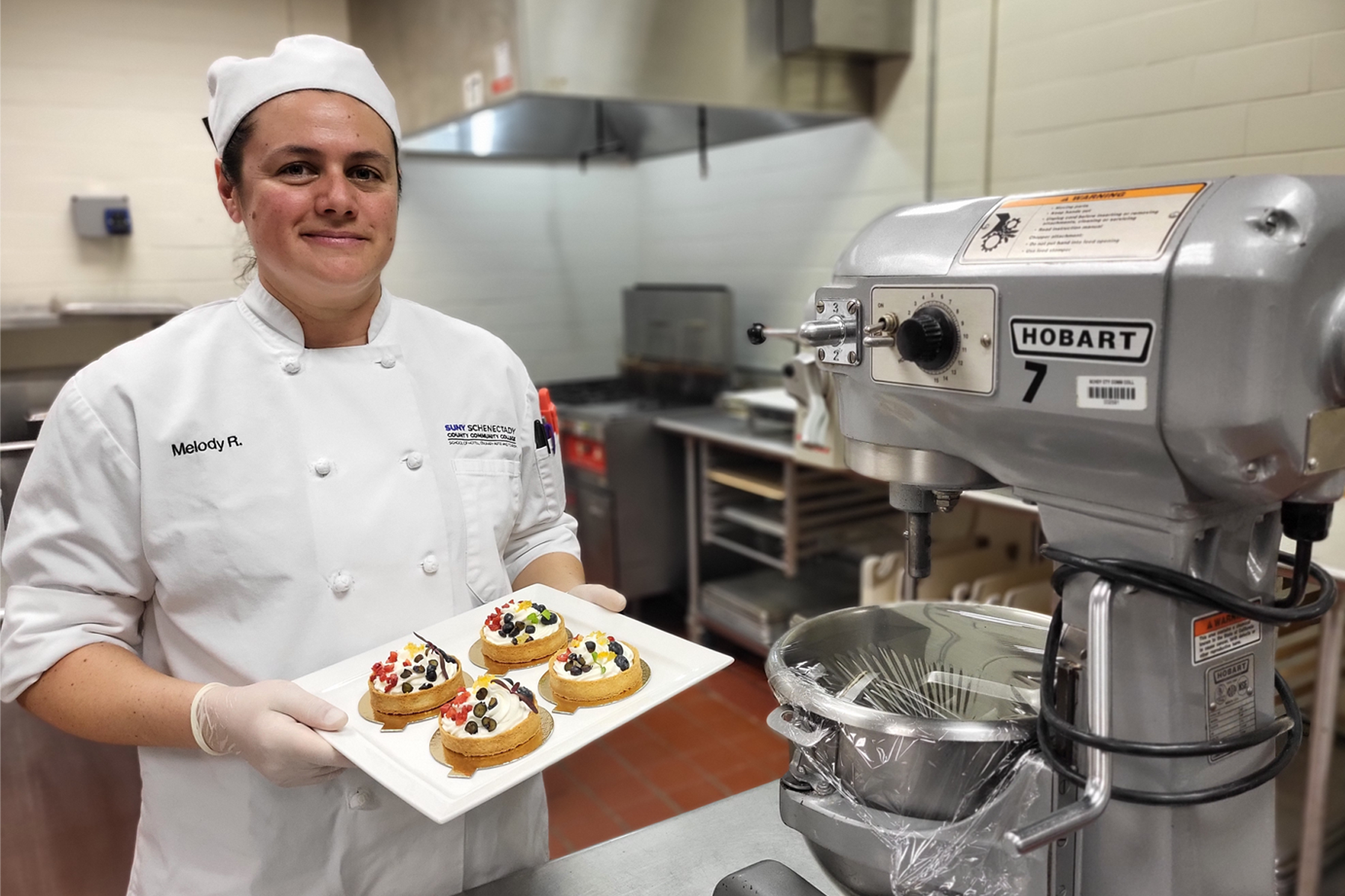 Melody Roper holding dish of baked goods, in culinary uniform, in culinary lab