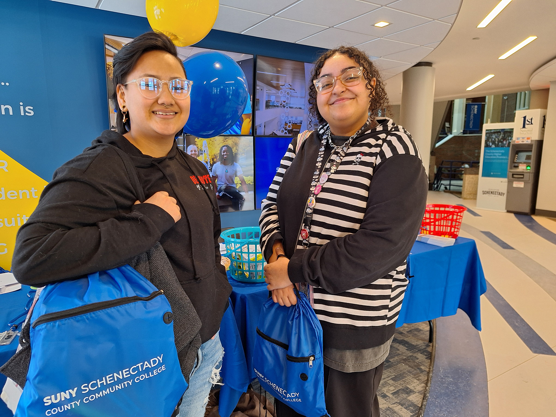 Two students, holding book bags with College name on them, standing, smiling