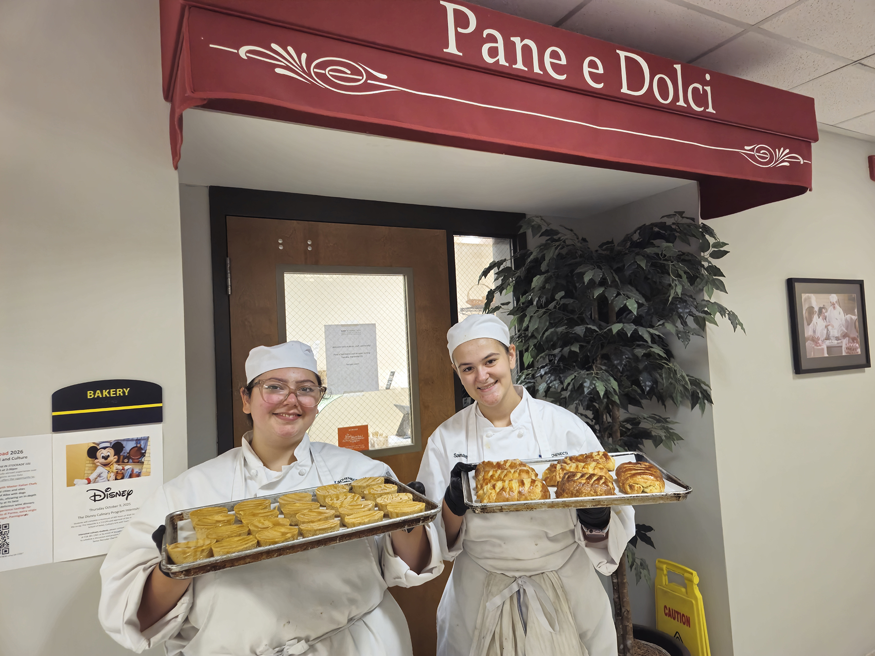 Two students holding trays of baked goods, standing in front of bakery