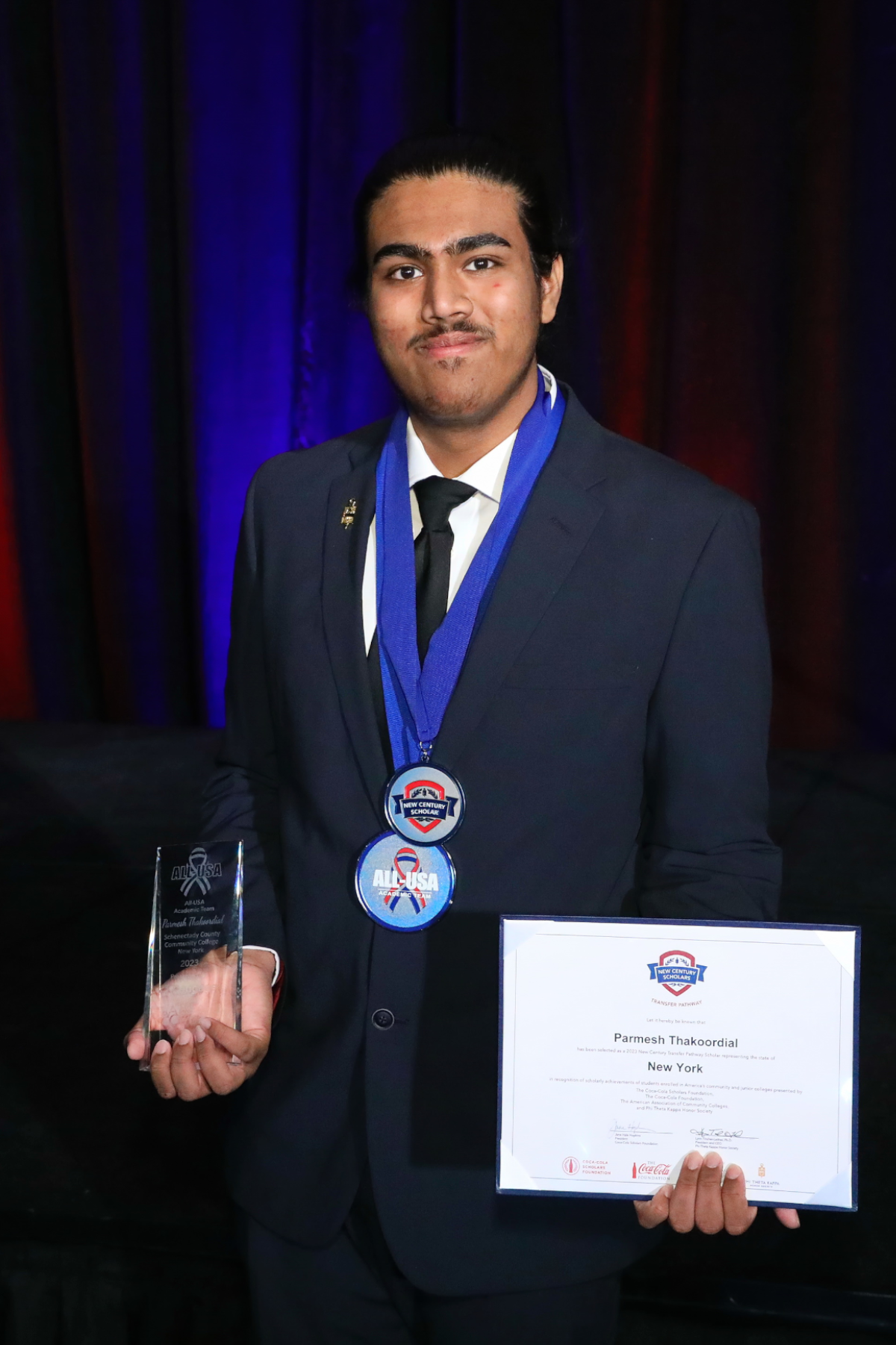 Parmesh Thakoordial smiling, holding awards for All-USA Academic Team and New Century Transfer Pathway Scholarship