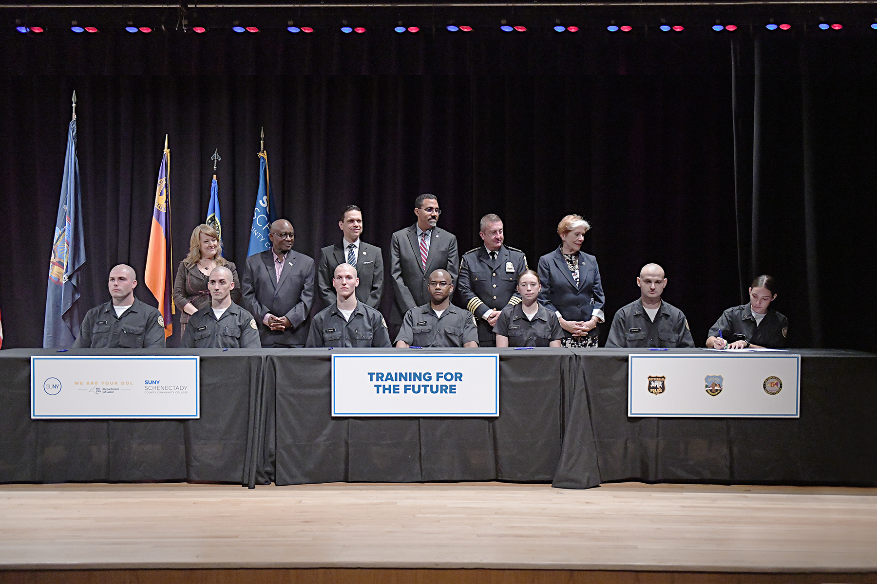 Police apprentice recruits sitting at table in auditorium signing, officials standing beind them