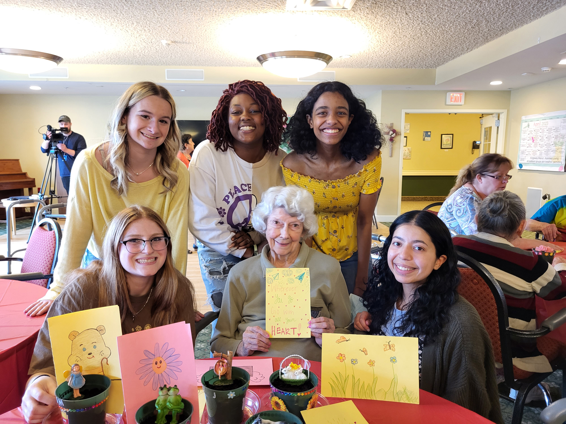 Students holding cards and flower pots with resident, seated at table