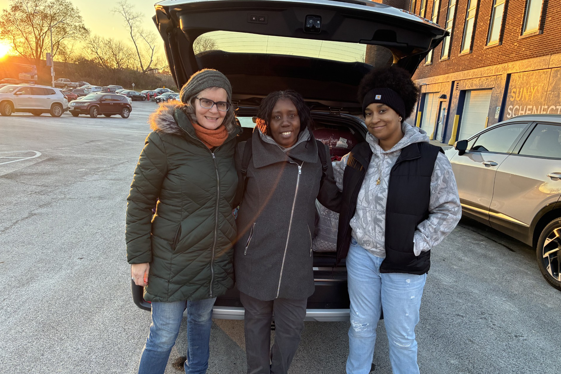 Students standing, smiling in front of car, trunk open with items they are donating