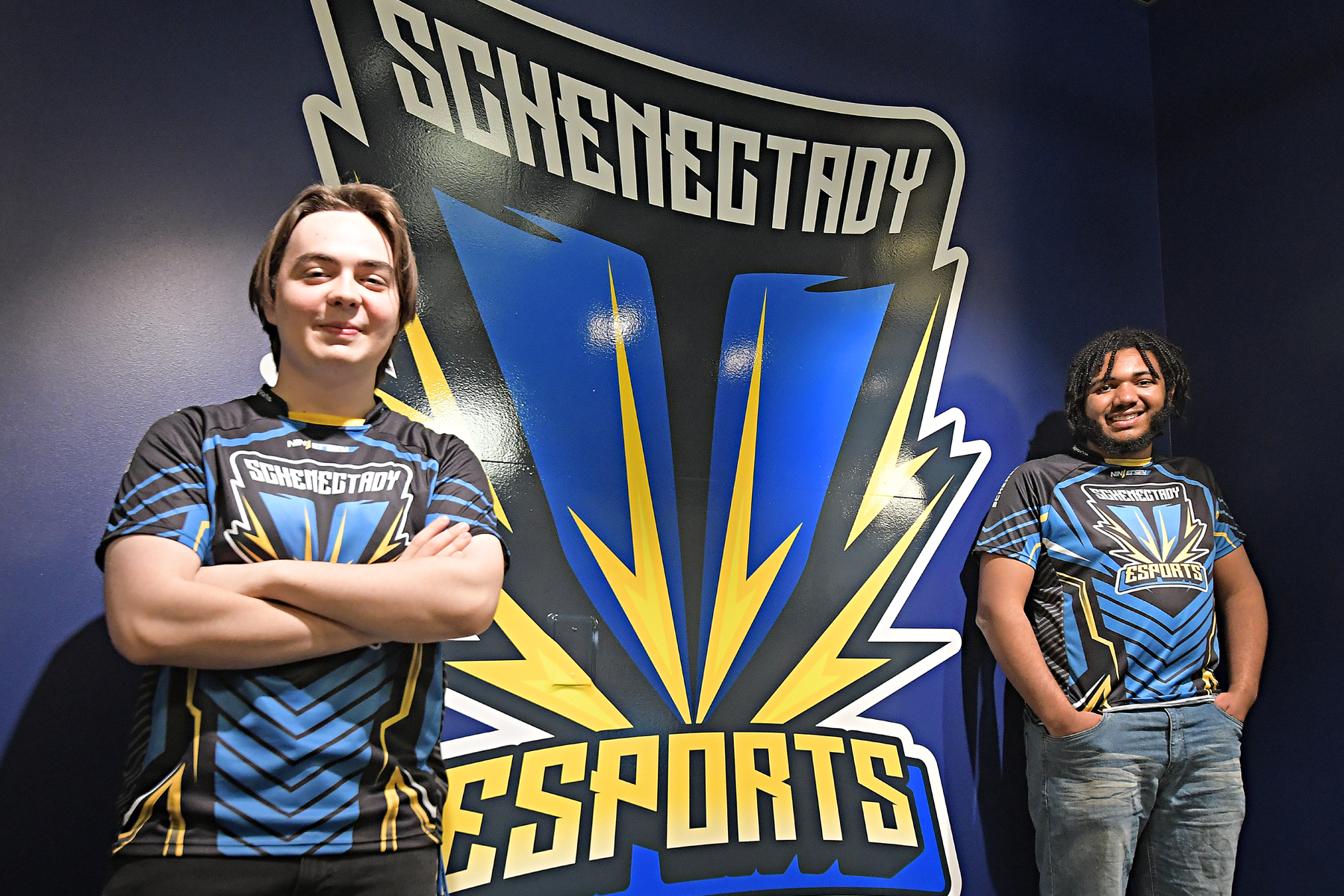 Two students, wearing eSports team jerseys and standing in front of eSports logo